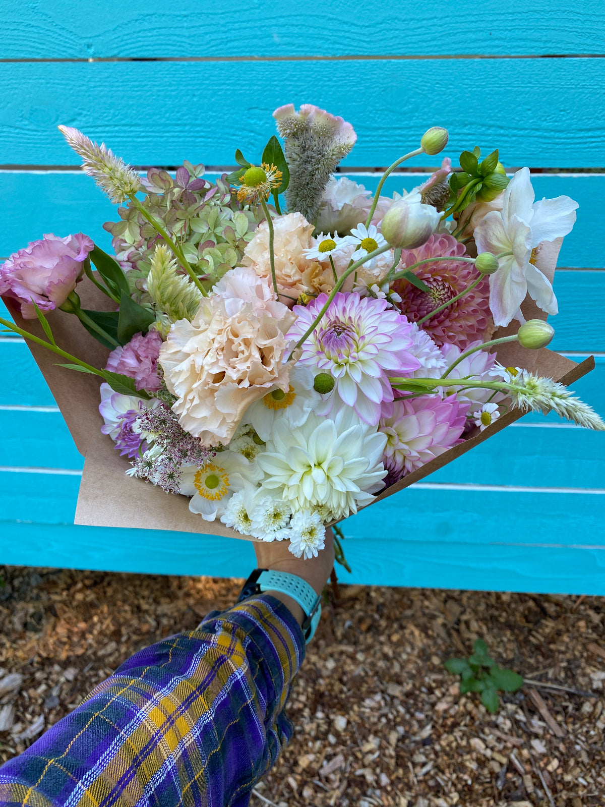 A hand holding a bouquet of mixed specialty cut flowers wrapped in brown kraft paper, set against a bright turquoise wooden fence and gravel ground.